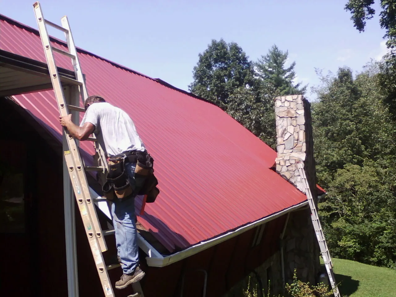 Roofer on a ladder beside a red metal roof and stone chimney — Soffit & Fascia Repair by Roofing Brothers in Watkinsville